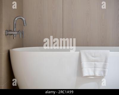 Neatly folded white towel rests on the edge of the white luxury curved ceramic bathtub near the steel faucet on light brown wooden wall background wit Stock Photo