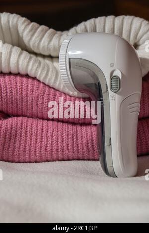 Anti-pilling razor. Device for shaving clothes. Anti-Plush fabric Shaver. Electric portable sweater pill defuzzer Lint remover from pink acrylic or wool sweater. Electric device fabric shaver after cleaning and collected fluff lint. Closeup Pilled sweater. Old used clothes with pilling.  Stock Photo