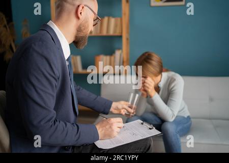 Depressed female patient sitting on psychotherapist couch as male doctor offers her a glass of water. Stock Photo