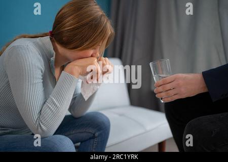 Depressed female patient sitting on psychotherapist couch as male doctor offers her a glass of water Stock Photo