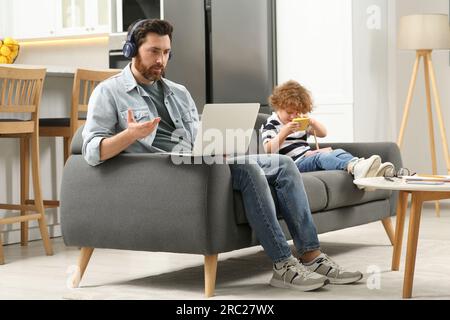 Father working remotely on laptop while his son playing with toys at home. Man having video chat with colleagues Stock Photo