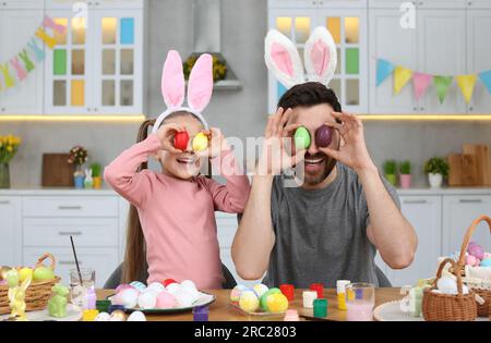 Father and his cute daughter covering eyes with beautifully painted Easter eggs at table in kitchen Stock Photo