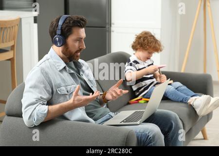 Father working remotely on laptop while his son playing with toys at home. Man having video chat with colleagues Stock Photo