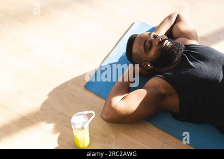 Relaxed african american man wearing sportswear with bottle of juice and lying on mat at home Stock Photo