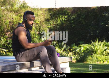 African american man wearing sportswear, headphones and using smartphone in garden Stock Photo
