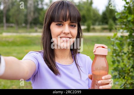 Cheerful smiling young female athlete takes photo selfie with smartphone and holding a water bottle after exercising outdoors in city park. Stock Photo