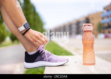 Sporty woman tying shoelaces on a bench at fitness training outdoors. Young female athlete running workout. Hydration, wellness, exercise, health. Stock Photo