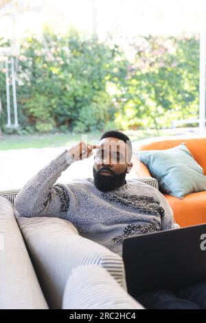 Thoughtful african american man wearing jumper and using laptop on couch at home Stock Photo
