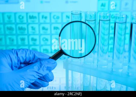 Scientist with magnifying glass examining test tubes against periodic table of elements in laboratory, closeup. Color tone effect Stock Photo