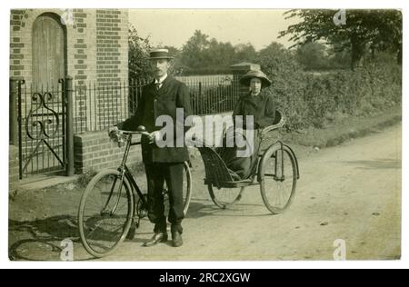 Early 1900's Edwardian era postcard of couple out cycling in the countryside, the wife is sitting in a trailer / bath chair (early wheelchair) attached to the bike whilst the husband does all the work. Early cargo bike! circa 1910, U.K. Stock Photo
