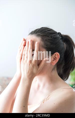Young adult Caucasian woman with hands covering her face. Conceptual image. Concept of physical or psychological pain or discomfort. Copy space Stock Photo