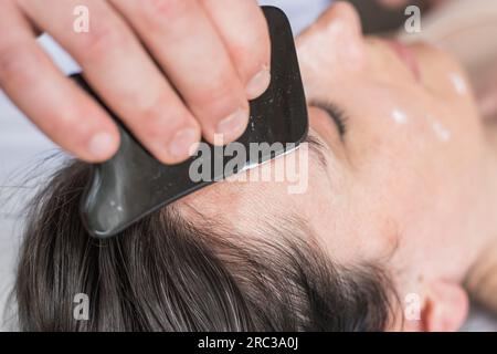 Young Caucasian woman getting facial treatment. Therapist doing skin scrape technique. Chinese technique, gua sha, for pain relief and treatment Stock Photo