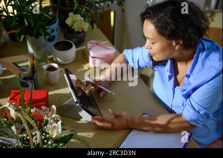 Overhead view of female florist, floral designer sketching on digital tablet, working on floral arrangement in a flower design studio, sitting at tabl Stock Photo