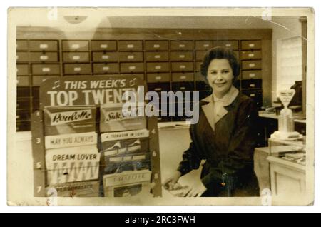 Original 1950's era photograph of an attractive, smiling female shop assistant inside a record store or general store with a record sales department, with This Week's Pop Top Twenty display stand, dated to 1959 from Ross Conway's Roulette which was no 1 in and his Sidesaddle hit also a number one hit in 1959, U.K. Stock Photo