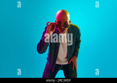 Portrait of young bald man with moustache wearing stylish clothes and sunglasses against blue studio background in neon light Stock Photo