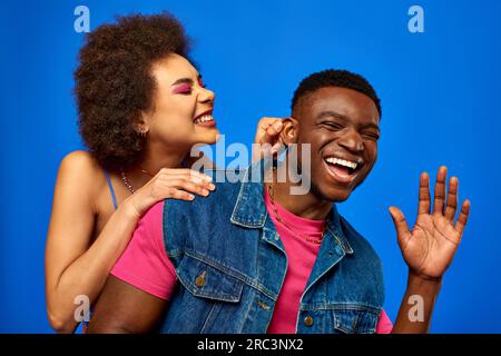 Positive young african american woman with bold makeup touching ear of stylish best friend in summer outfit while having fun and standing isolated on Stock Photo