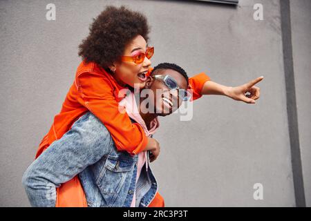 Excited young african american woman in sunglasses and stylish outfit pointing with finger while piggybacking on friend and standing near building on Stock Photo