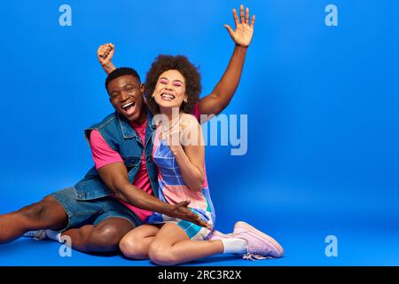 Cheerful young african american best friends in bright summer outfits waving hand and showing yes gesture at camera while sitting together on blue bac Stock Photo