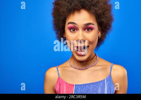 Portrait of stylish young african american woman with natural hair and bold makeup sticking out tongue and looking at camera while standing isolated o Stock Photo