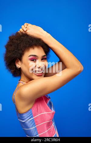 Portrait of pleased young african american woman with bold makeup wearing summer sundress while posing and looking at camera isolated on blue, charism Stock Photo