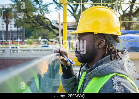 profile of male engineer of african ethnicity with dreadlocks and beard, standing outdoors with helmet and safety vest supervising the construction si Stock Photo
