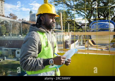 portrait of young black man with beard and dreadlocks engineer dressed in jacket and wearing vest and safety helmet, is standing outdoors holding fold Stock Photo