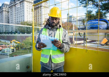 front view of young black engineer man in yellow safety helmet, green vest, foreman of construction site standing outdoors reading documents with inst Stock Photo