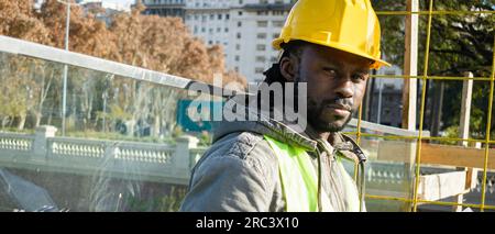 banner of young black engineer man with beard, wearing safety helmet and vest, wearing jacket, is standing outdoors at construction site looking at ca Stock Photo