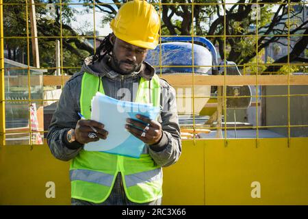 black ethnic male civil engineer wearing yellow hardhat, green jacket and vest, standing at construction site, reading a folder, copy space. Stock Photo