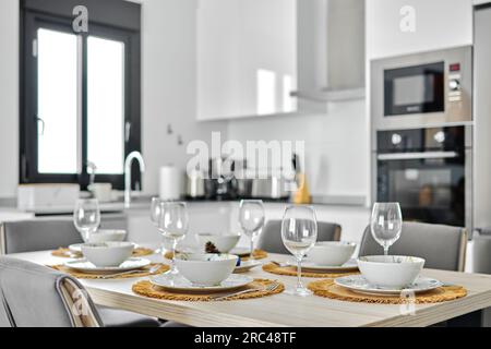 In modern fashionable cozy kitchen empty wine glasses and dinnerware is ready for lunch served on dining table, no people Stock Photo