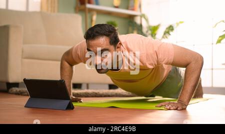 Indian man doing push ups exercise by online video classes from tablet at home - concept of Fitness instruction, Exercise Tutorial and Bodyweight Stock Photo