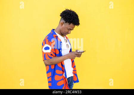 Side view of casually dressed young African American male with curly hair hairstyle browsing smartphone while standing against yellow background Stock Photo