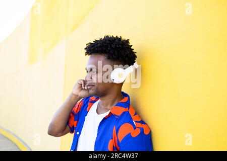 Thoughtful African American male in colorful shirt listening to music from wireless headphones while standing looking away against yellow wall backgro Stock Photo