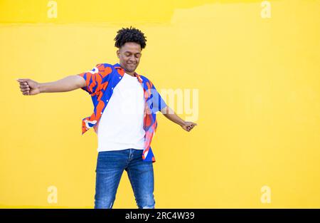 Cheerful American male with dreadlocks and in colorful shirt dancing while swinging hands against yellow background Stock Photo