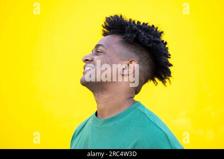 Positive African American male with curly hair hair and trendy casual clothes smiling looking away while standing on yellow background Stock Photo