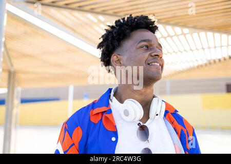 Portrait of confident African American male with curly hair and headphones smiling and looking away while standing under shining roof on city street Stock Photo