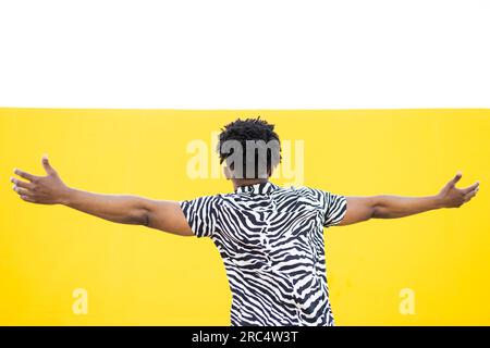 Back view of unrecognizable African American male in trendy zebra shirt smiling with raised head and stretched hands while dancing against yellow back Stock Photo