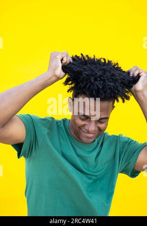 Cheerful young African American male in curly hair and casual clothes with raised hands touching hair while standing on yellow background and looking Stock Photo