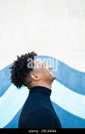Side view of positive young African American male with curly hair hairstyle in casual clothes standing against blue and white wall while looking up Stock Photo