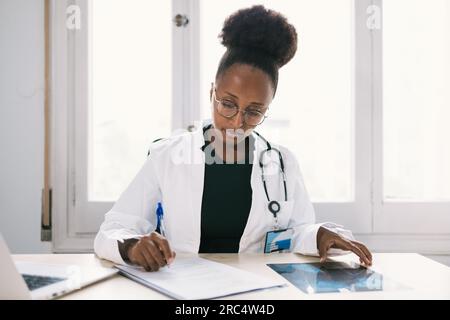 Ethnic female physician with stethoscope sitting at desk and focusing while taking notes in modern hospital Stock Photo