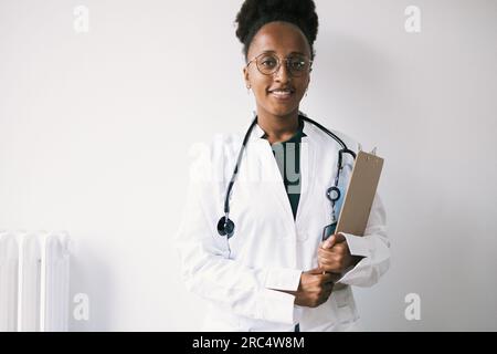 Portrait of positive young African American female doctor in eyeglasses, uniform and stethoscope smiling and looking at camera while standing with cli Stock Photo