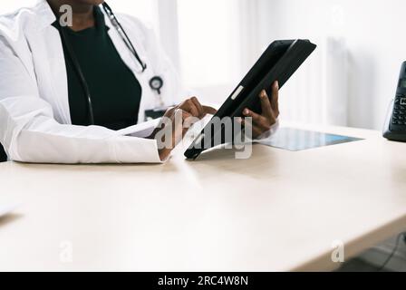 Crop African American female doctor in uniform and stethoscope with tablet while sitting at table in daylight Stock Photo