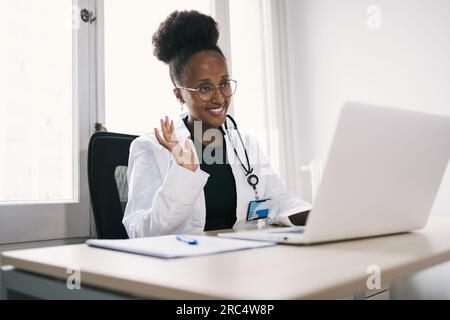 Positive African American female doctor in medical uniform waving at laptop during online consultation in clinic Stock Photo