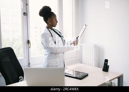 Side view of concentrated African American female doctor in uniform with stethoscope looking away while standing near table with laptop and tablet and Stock Photo