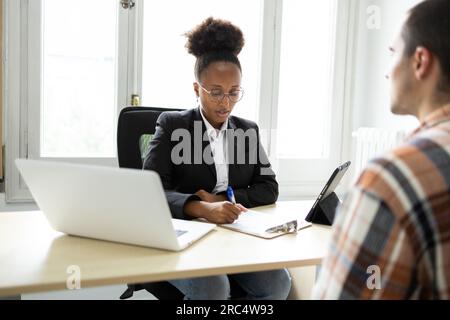 Focused young African American female lawyer in eyeglasses looking down at notes on clipboard while sitting at table with laptop, tablet and interacti Stock Photo
