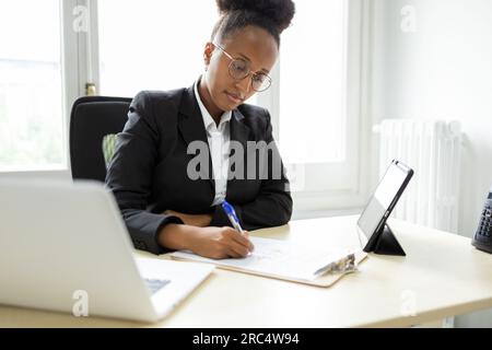 Concentrated young African American businesswoman in formal clothes, eyeglasses looking down and writing notes on clipboard while sitting at table wit Stock Photo