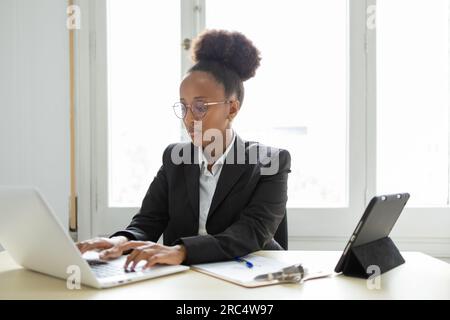 Concentrated young African American female entrepreneur in elegant suit and eyeglasses looking at screen of laptop while sitting at table with clipboa Stock Photo
