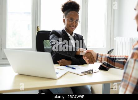 Smiling young African American female executive in jacket and eyeglasses shaking hands with anonymous male client and looking at each other while sitt Stock Photo