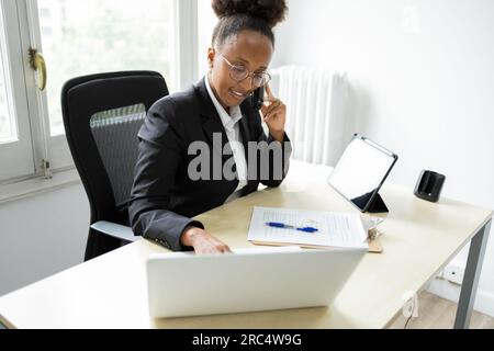 High angle of young African American female bank manager speaking on phone and looking down while typing on laptop and sitting at table with tablet an Stock Photo
