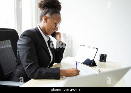 Side view of young African American female bank manager speaking on phone and looking down while writing notes and sitting at table with tablet in off Stock Photo
